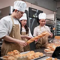 Un chef boulanger montre comment façonner des baguettes à deux apprenties en cuisine.